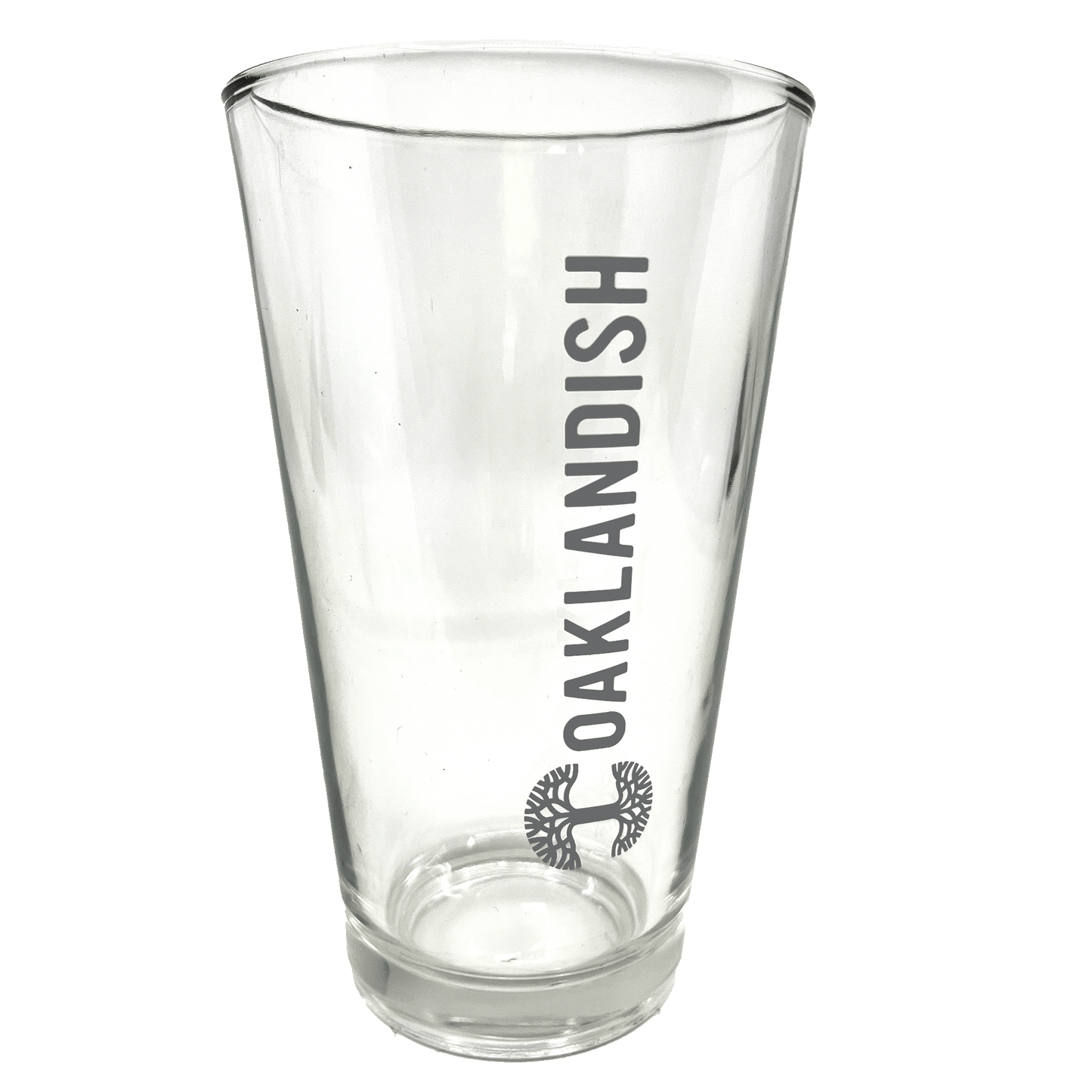 Image of the Oaklandish Banner Pint Glass—a clear, tapered drinkware item featuring the dark grey Oaklandish logo printed vertically. Below the logo is a black image of a spreading oak tree. The background is entirely white, emphasizing the pint glass.