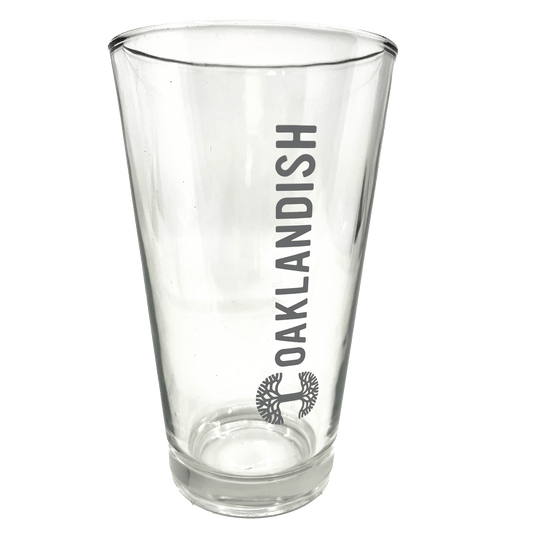 Image of the Oaklandish Banner Pint Glass—a clear, tapered drinkware item featuring the dark grey Oaklandish logo printed vertically. Below the logo is a black image of a spreading oak tree. The background is entirely white, emphasizing the pint glass.