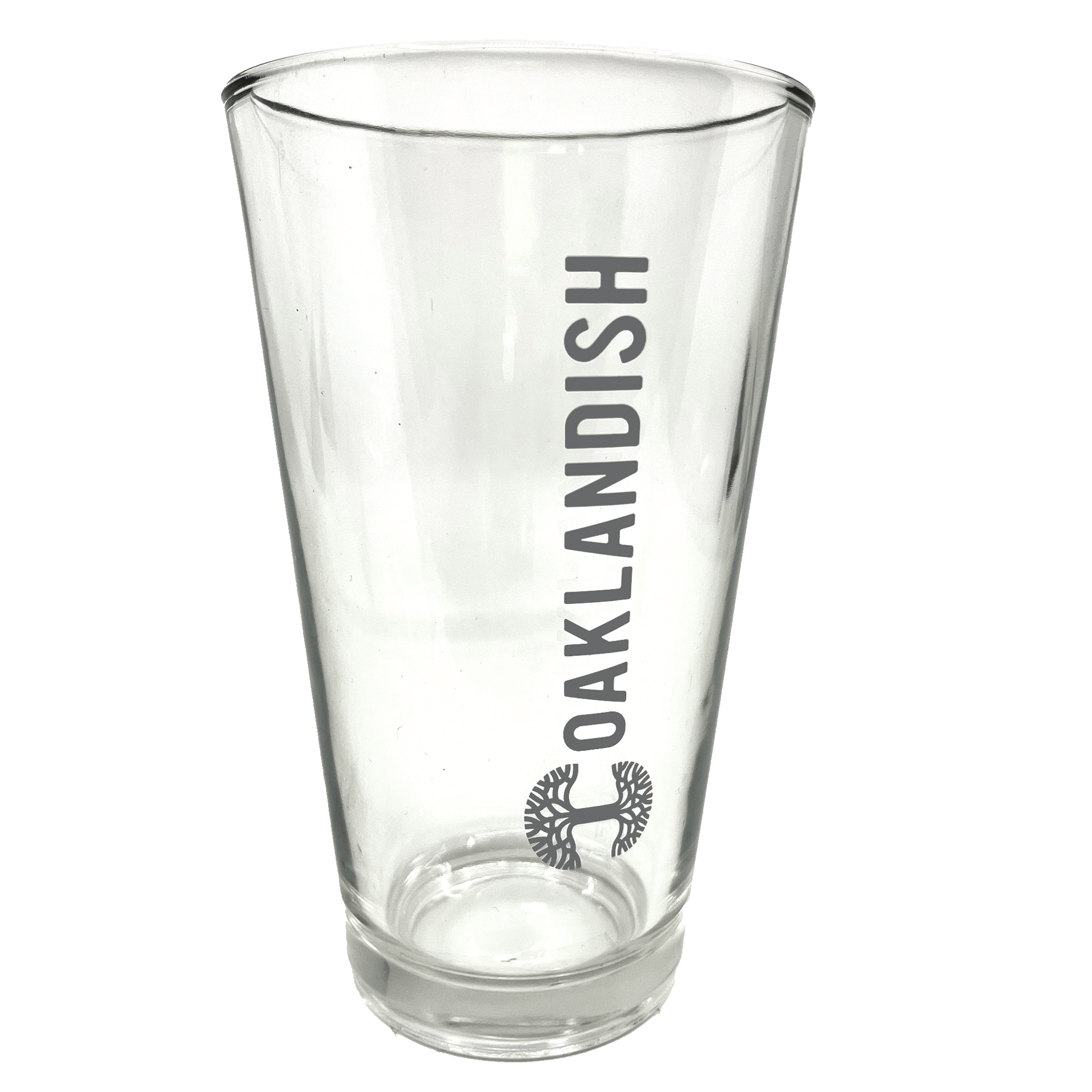 Image of the Oaklandish Banner Pint Glass—a clear, tapered drinkware item featuring the dark grey Oaklandish logo printed vertically. Below the logo is a black image of a spreading oak tree. The background is entirely white, emphasizing the pint glass.