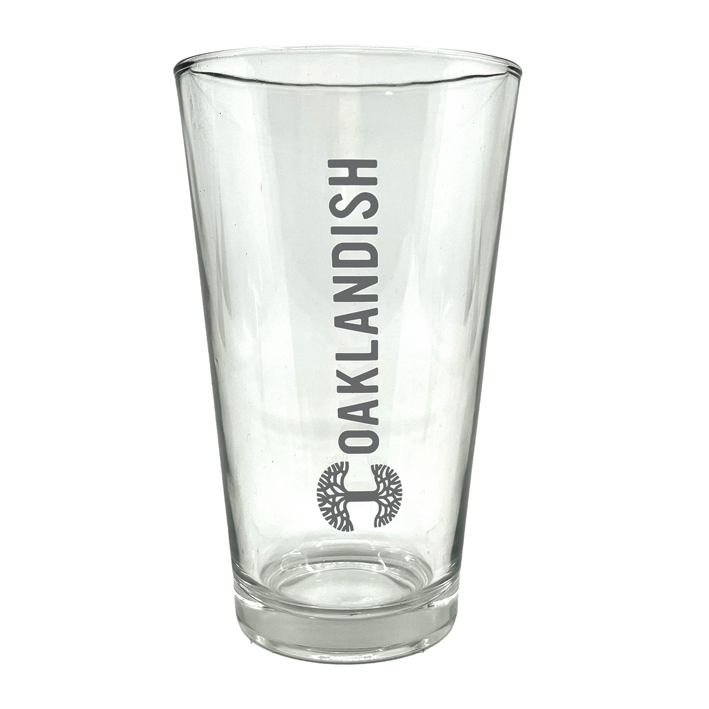 The Banner Pint Glass by Oaklandish is a clear, tall pint glass featuring a bold, dark grey Oaklandish logo in vertical text centered on one side. Below the text is a stylized black tree design within a circular outline. This piece of drinkware boasts a smooth finish and remains empty inside, awaiting your favorite beverage.