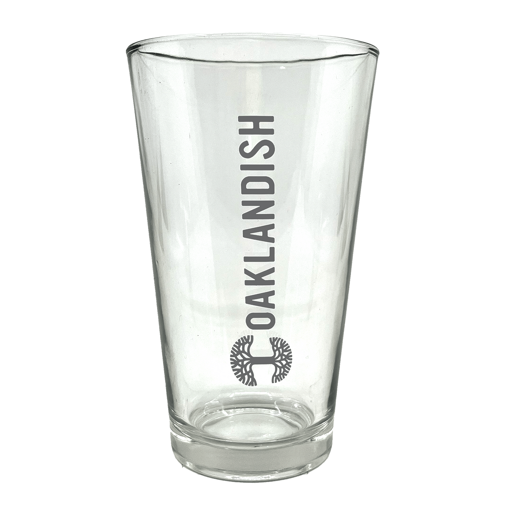 The Banner Pint Glass by Oaklandish is a clear, tall pint glass featuring a bold, dark grey Oaklandish logo in vertical text centered on one side. Below the text is a stylized black tree design within a circular outline. This piece of drinkware boasts a smooth finish and remains empty inside, awaiting your favorite beverage.