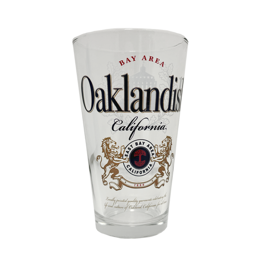 The Lager Pint Glass by Oaklandish is a 16 oz pint glass featuring the text "Bay Area Oaklandish California" in black and red. Below this, intricate graphics depict two lions standing on their hind legs, flanking a red and black emblem that reads "East Bay" and "California." Additional small text appears beneath the emblem, making it perfect for any drinkware collection.