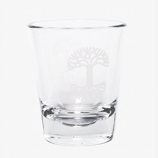 The Oaklandish Trees Shot Glass by Oaklandish is a clear glass tumbler with a thick, solid base, showcasing an elegant logo etching of a stylized tree with sprawling branches and roots, symbolizing the tree of life. This exquisite shot glass is empty and set against a plain white background.